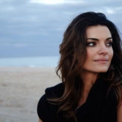 Mulher de cabelos castanhos olha para a lateral em uma praia de areia clara, com mar e céu nublado ao fundo.