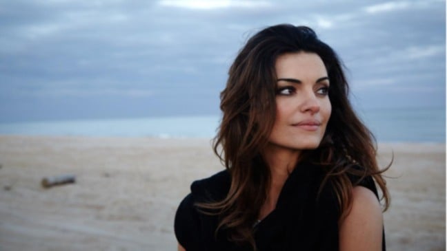 Mulher de cabelos castanhos olha para a lateral em uma praia de areia clara, com mar e céu nublado ao fundo.