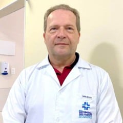 Médico do Hospital São Vicente, Dr. Eduardo Leme, em corredor da unidade, usando jaleco branco e olhando para a câmera.