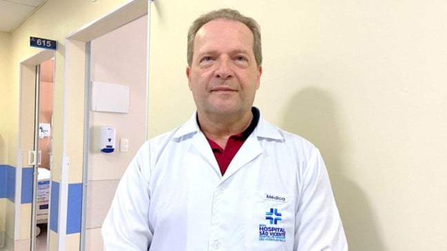Médico do Hospital São Vicente, Dr. Eduardo Leme, em corredor da unidade, usando jaleco branco e olhando para a câmera.