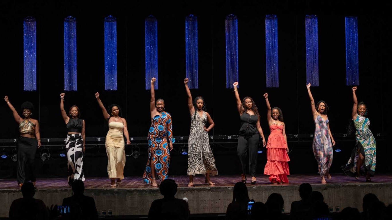 Candidatas da Miss Pérola Negra 2025 alinhadas no palco do Teatro Polytheama, todas com o braço levantado em gesto de empoderamento, sob iluminação azul. Candidatas da Miss Pérola Negra 2025 alinhadas no palco do Teatro Polytheama, todas com o braço levantado em gesto de empoderamento, sob iluminação azul.
