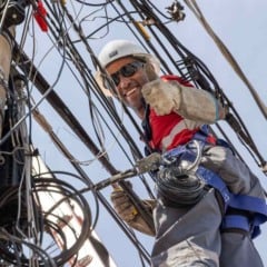 Técnico de capacete e equipamentos de segurança organiza cabos em um poste cheio de fiações e faz sinal de positivo para a câmera.