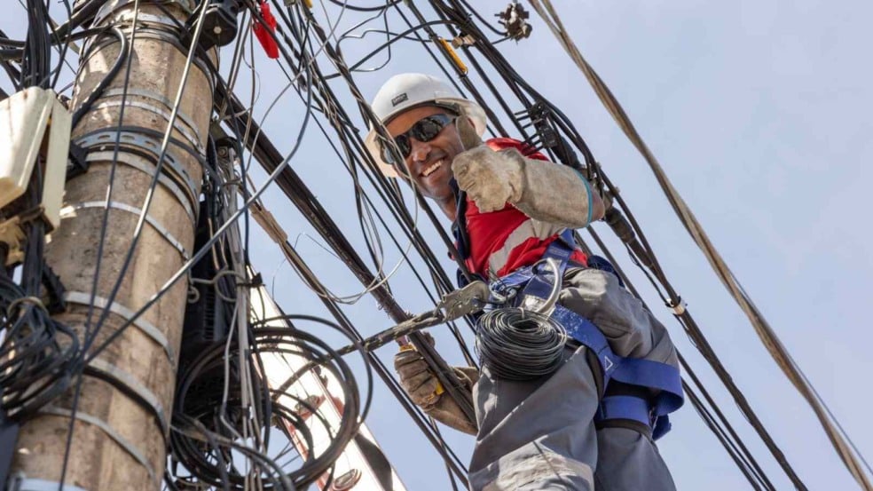 Técnico de capacete e equipamentos de segurança organiza cabos em um poste cheio de fiações e faz sinal de positivo para a câmera. Técnico de capacete e equipamentos de segurança organiza cabos em um poste cheio de fiações e faz sinal de positivo para a câmera.