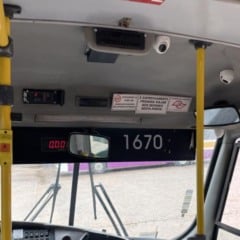 Interior de um ônibus escolar com câmeras de videomonitoramento instaladas no teto, avisos de segurança, barras de apoio amarelas e adesivo informando que o passageiro está sendo filmado.