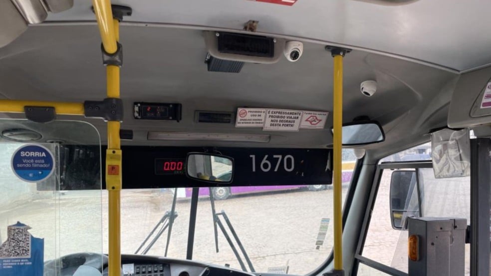 Interior de um ônibus escolar com câmeras de videomonitoramento instaladas no teto, avisos de segurança, barras de apoio amarelas e adesivo informando que o passageiro está sendo filmado.