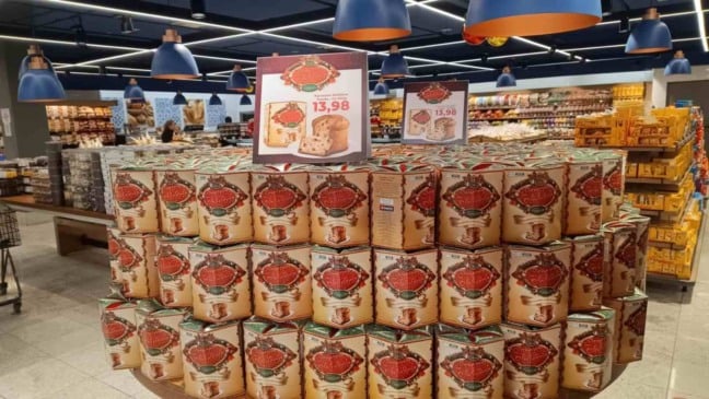 Empilhamento de panetones da campanha Panetone Solidário Tauste com cartaz exibindo o preço de R$ 13,98 dentro de um supermercado.