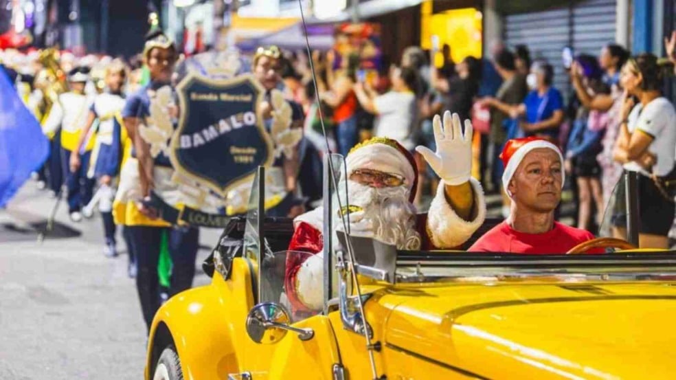 Papai Noel acena de dentro de um carro conversível amarelo durante um desfile, com banda marcial e público acompanhando o evento nas laterais da rua.