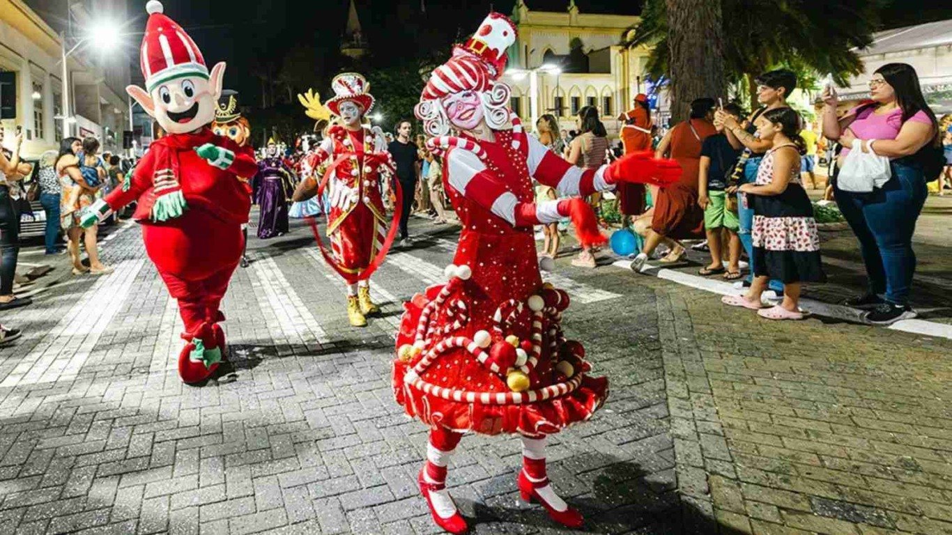 Cena de um desfile natalino em rua iluminada, com artistas fantasiados — incluindo um duende e uma personagem com vestido decorado como doces — caminhando e interagindo com o público, enquanto moradores observam e fotografam a apresentação.