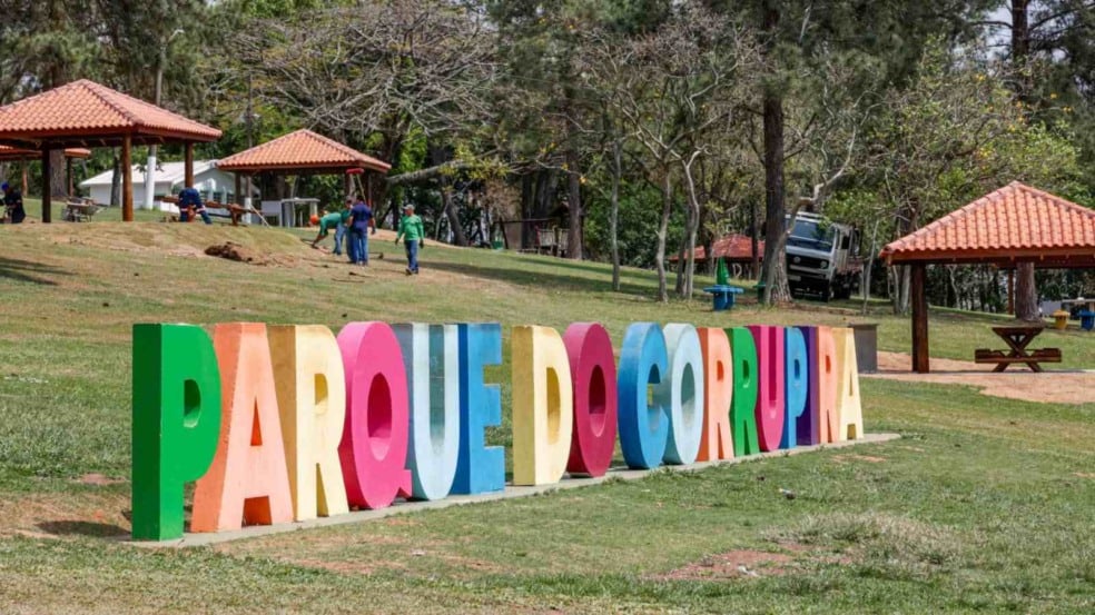 Letreiro colorido com a frase “Parque do Corrupira” em área gramada, com quiosques, árvores e visitantes ao fundo em um parque de Jundiaí. Letreiro colorido com a frase “Parque do Corrupira” em área gramada, com quiosques, árvores e visitantes ao fundo em um parque de Jundiaí.