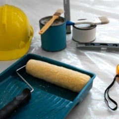 Materiais de pintura predial, incluindo rolo, pincéis, capacete de segurança, latas de tinta, trena e nível, dispostos sobre uma superfície coberta por plástico.