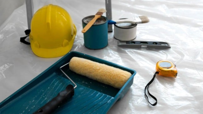 Materiais de pintura predial, incluindo rolo, pincéis, capacete de segurança, latas de tinta, trena e nível, dispostos sobre uma superfície coberta por plástico.