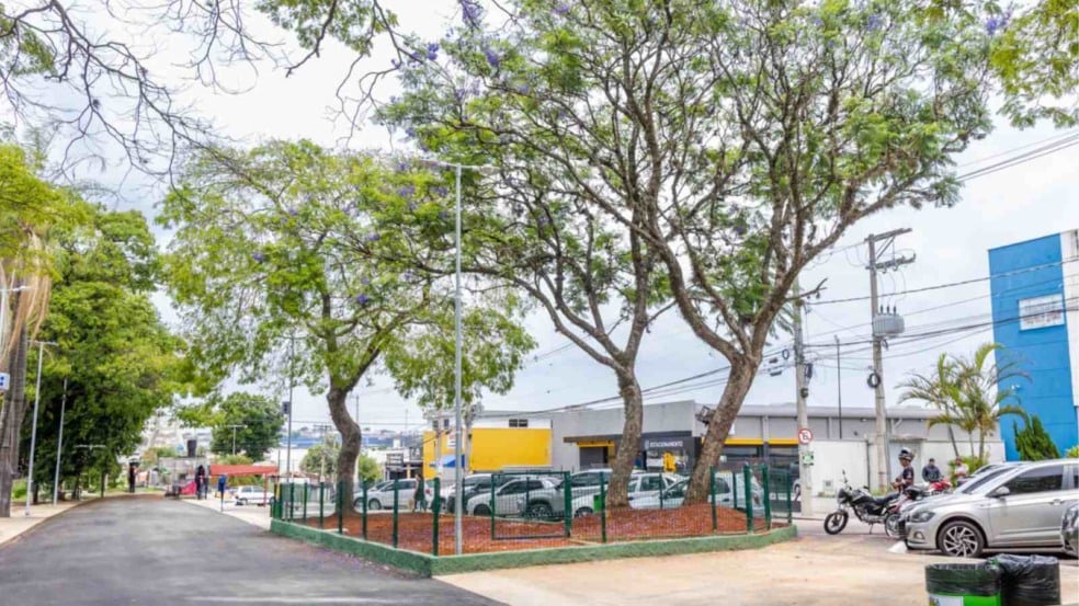 Área revitalizada da Praça Joaquim Soares Lemos em Jundiaí, com árvores floridas, via asfaltada, espaço cercado e veículos estacionados ao lado.