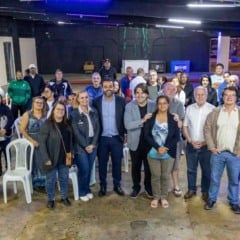 Foto mostra o prefeito de Jundiaí, Gustavo Martinelli, posando ao centro com um grupo de moradores e representantes da Prefeitura em um salão amplo. Pessoas de diferentes idades estão sorrindo para a câmera, demonstrando participação no encontro sobre regularização fundiária da Vila Arens.
