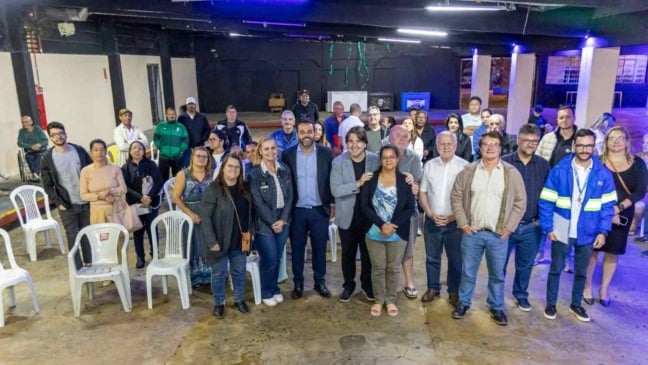 Foto mostra o prefeito de Jundiaí, Gustavo Martinelli, posando ao centro com um grupo de moradores e representantes da Prefeitura em um salão amplo. Pessoas de diferentes idades estão sorrindo para a câmera, demonstrando participação no encontro sobre regularização fundiária da Vila Arens.
