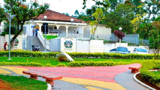 Fachada da Prefeitura Municipal de Cajamar com jardim arborizado, escadaria de acesso e pessoas circulando na entrada do prédio.