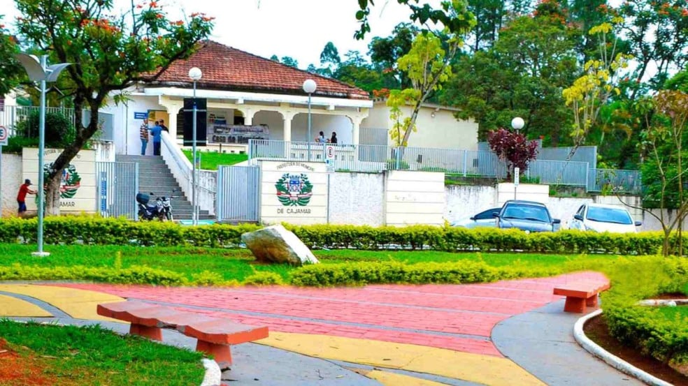 Fachada da Prefeitura Municipal de Cajamar com jardim arborizado, escadaria de acesso e pessoas circulando na entrada do prédio.