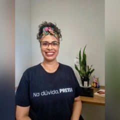 Mulher sorrindo em ambiente interno, usando óculos, lenço colorido no cabelo e camiseta preta com a frase “Na dúvida, PRETO!”. Ao fundo, há uma mesa com uma planta e itens de escritório.