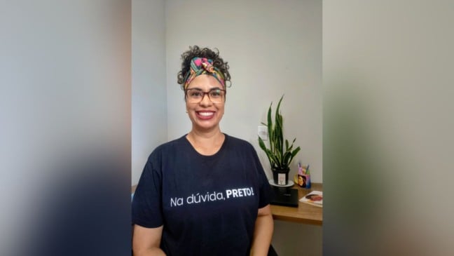 Mulher sorrindo em ambiente interno, usando óculos, lenço colorido no cabelo e camiseta preta com a frase “Na dúvida, PRETO!”. Ao fundo, há uma mesa com uma planta e itens de escritório.