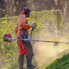 Pessoa usando uniforme laranja, máscara e equipamentos de segurança opera uma roçadeira para cortar mato em área verde ao ar livre.