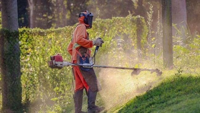 Pessoa usando uniforme laranja, máscara e equipamentos de segurança opera uma roçadeira para cortar mato em área verde ao ar livre.
