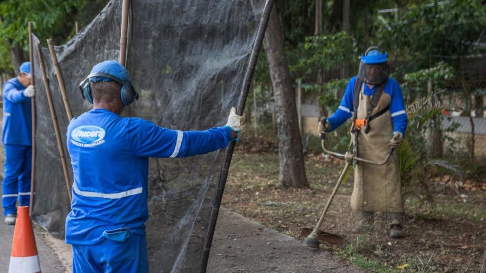 Funcionários com uniformes azuis utilizam tela de proteção enquanto um trabalhador opera uma roçadeira durante serviço de limpeza e roçada em área verde de Jundiaí.