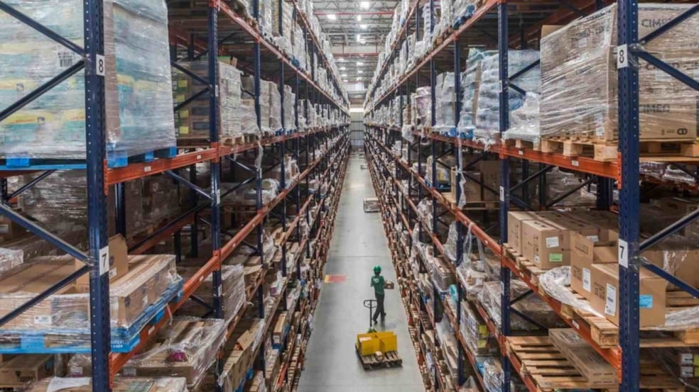 Centro de distribuição com grandes prateleiras cheias de mercadorias e funcionário operando paleteira no corredor.
