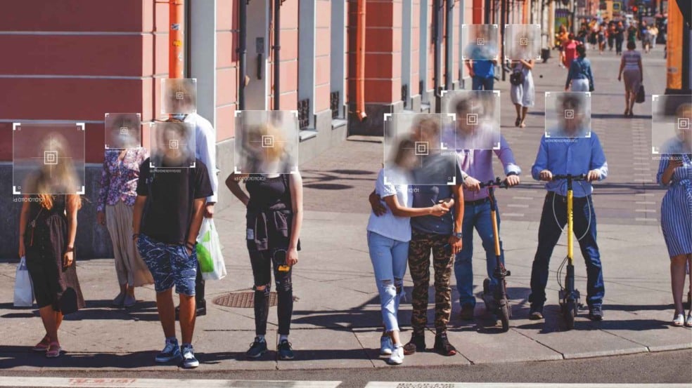 Grupo de pedestres em uma rua movimentada com seus rostos desfocados e marcadores digitais simulando reconhecimento facial, incluindo pessoas caminhando e duas usando patinetes elétricos. Grupo de pedestres em uma rua movimentada com seus rostos desfocados e marcadores digitais simulando reconhecimento facial, incluindo pessoas caminhando e duas usando patinetes elétricos.