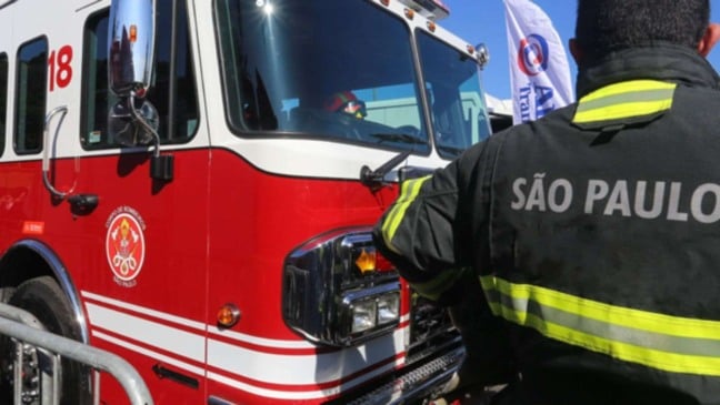 Bombeiro de São Paulo ao lado de caminhão vermelho do Corpo de Bombeiros em dia ensolarado.