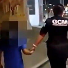 Guarda civil municipal caminha à noite de mãos dadas com uma criança com o rosto desfocado, acompanhando o menino pela calçada de uma rua iluminada.