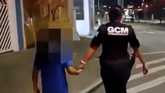 Guarda civil municipal caminha à noite de mãos dadas com uma criança com o rosto desfocado, acompanhando o menino pela calçada de uma rua iluminada.