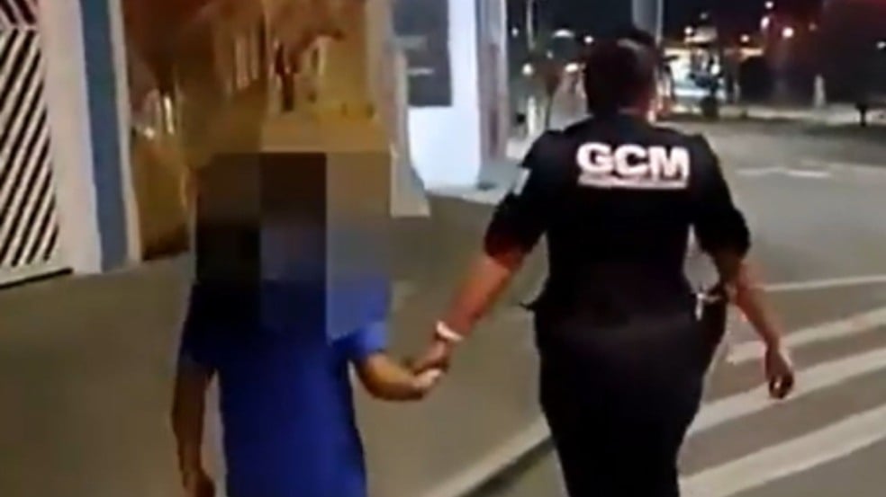 Guarda civil municipal caminha à noite de mãos dadas com uma criança com o rosto desfocado, acompanhando o menino pela calçada de uma rua iluminada. Guarda civil municipal caminha à noite de mãos dadas com uma criança com o rosto desfocado, acompanhando o menino pela calçada de uma rua iluminada.