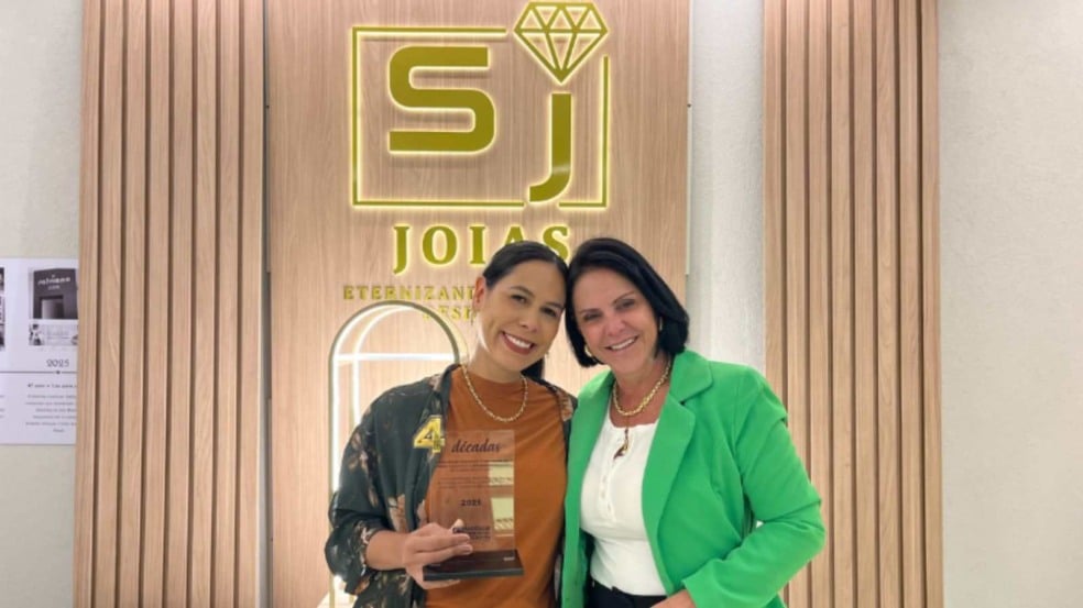 Talita e Maria Salviano posam sorrindo na loja da Salviano Jóias; Talita segura a placa de homenagem da ACE Jundiaí, com o logotipo iluminado da joalheria ao fundo.