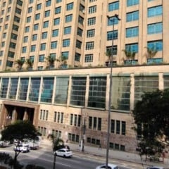 Imagem da sede da Secretaria da Fazenda de São Paulo, prédio alto com janelas amplas, terraço com plantas e movimento de carros na avenida em frente.