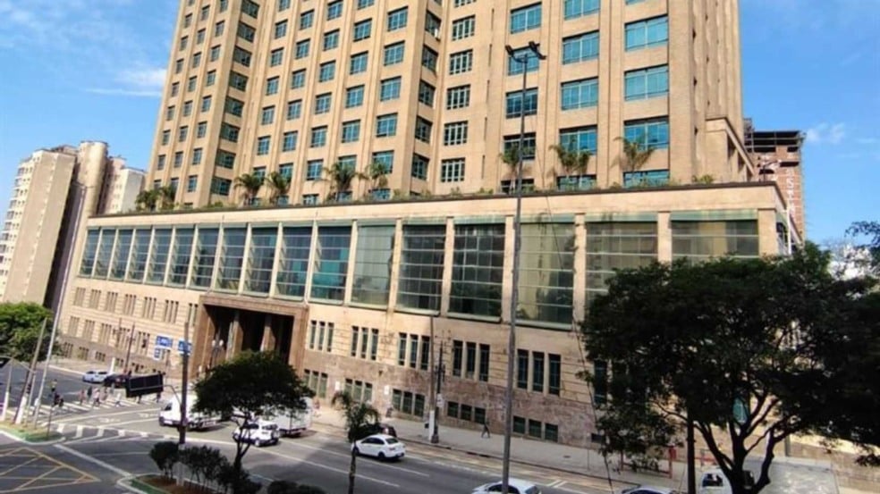 Imagem da sede da Secretaria da Fazenda de São Paulo, prédio alto com janelas amplas, terraço com plantas e movimento de carros na avenida em frente.