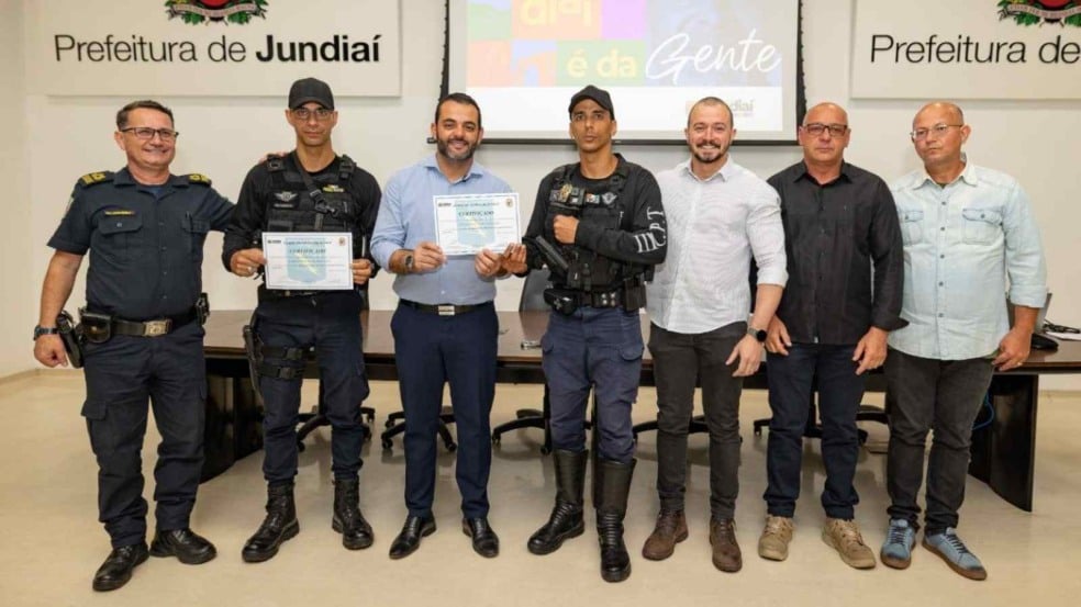 Grupo formado por guardas municipais e representantes da Prefeitura de Jundiaí posando para foto oficial; dois agentes seguram certificados recebidos durante a cerimônia. Grupo formado por guardas municipais e representantes da Prefeitura de Jundiaí posando para foto oficial; dois agentes seguram certificados recebidos durante a cerimônia.