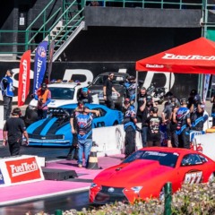 Carros de arrancada vermelho e azul alinhados na pista do SPID, com equipes técnicas nos boxes, bandeiras de patrocinadores e estrutura da FuelTech ao fundo durante o evento.