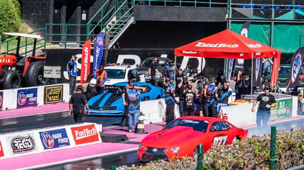 Carros de arrancada vermelho e azul alinhados na pista do SPID, com equipes técnicas nos boxes, bandeiras de patrocinadores e estrutura da FuelTech ao fundo durante o evento. Carros de arrancada vermelho e azul alinhados na pista do SPID, com equipes técnicas nos boxes, bandeiras de patrocinadores e estrutura da FuelTech ao fundo durante o evento.