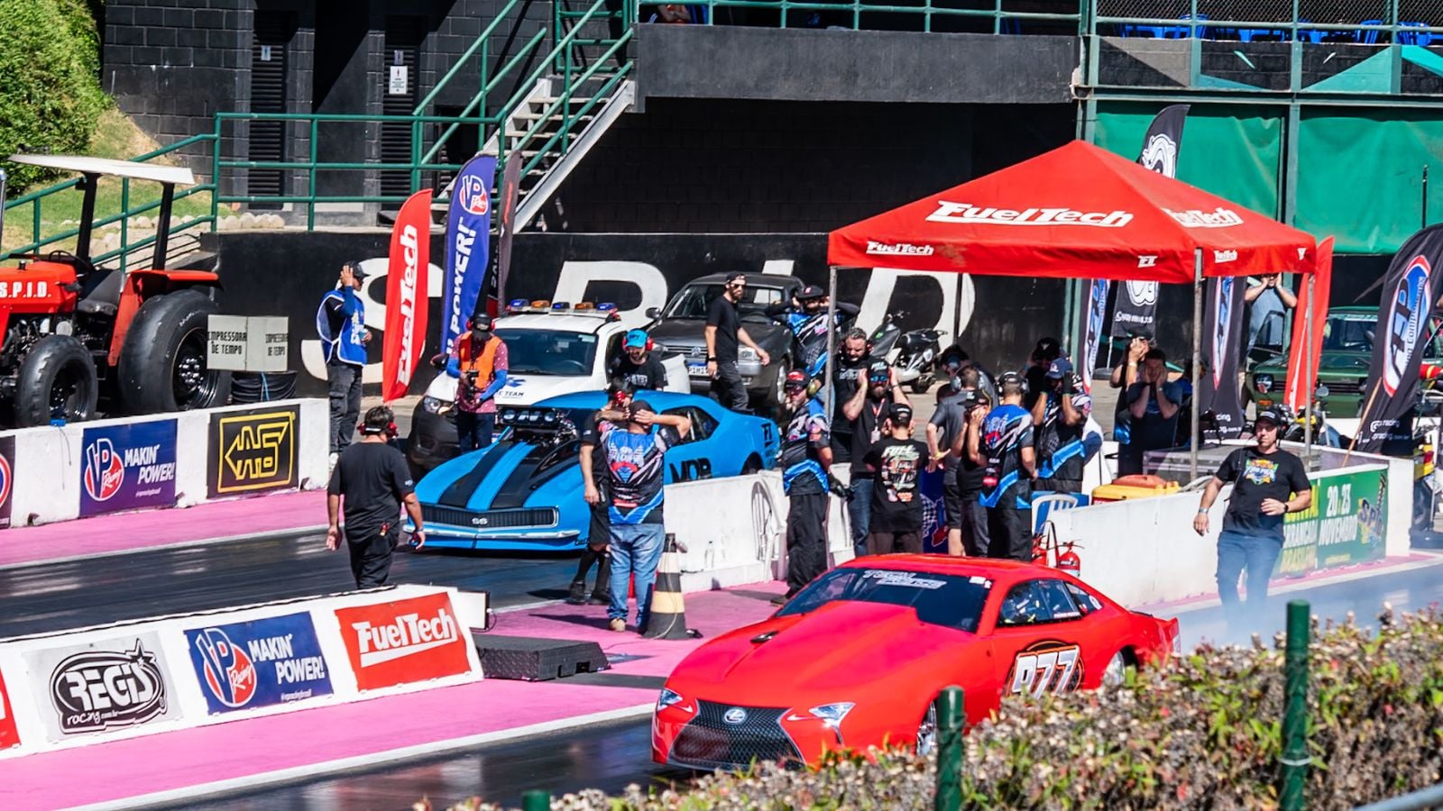 Carros de arrancada vermelho e azul alinhados na pista do SPID, com equipes técnicas nos boxes, bandeiras de patrocinadores e estrutura da FuelTech ao fundo durante o evento.