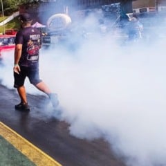Carro vermelho fazendo burnout e produzindo muita fumaça na pista do SPID Dragway, enquanto um membro da equipe caminha ao lado durante a preparação para a largada.