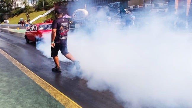 Carro vermelho fazendo burnout e produzindo muita fumaça na pista do SPID Dragway, enquanto um membro da equipe caminha ao lado durante a preparação para a largada.
