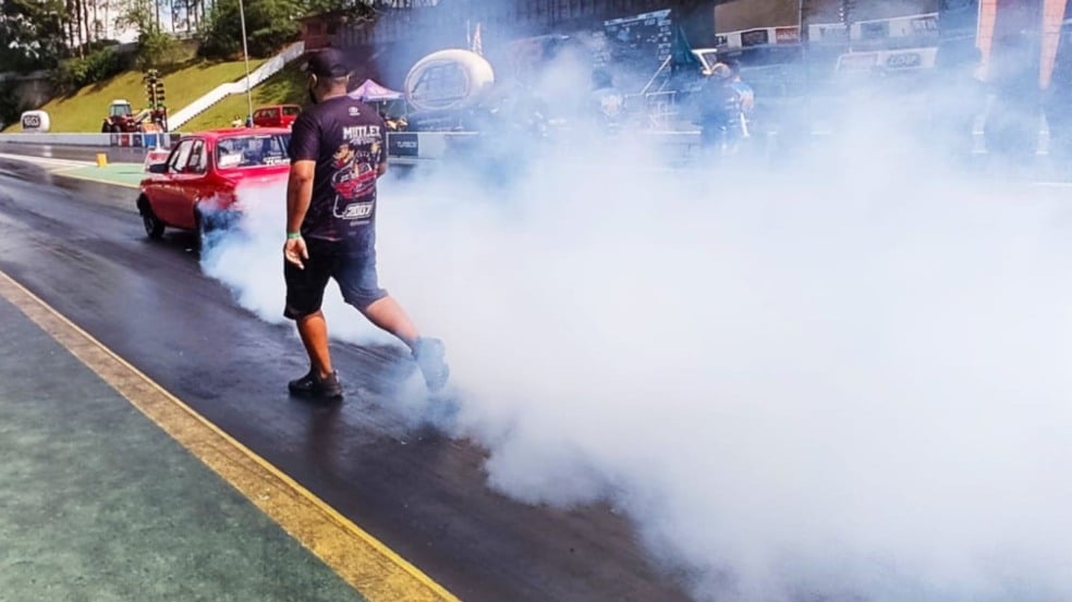 Carro vermelho fazendo burnout e produzindo muita fumaça na pista do SPID Dragway, enquanto um membro da equipe caminha ao lado durante a preparação para a largada.