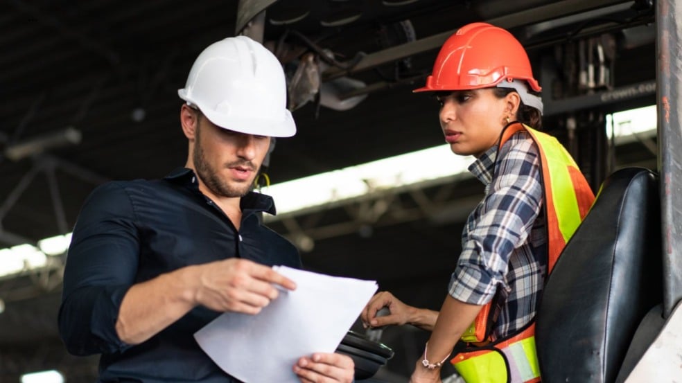 Profissionais com capacetes de segurança analisam um documento em galpão industrial. Profissionais com capacetes de segurança analisam um documento em galpão industrial.