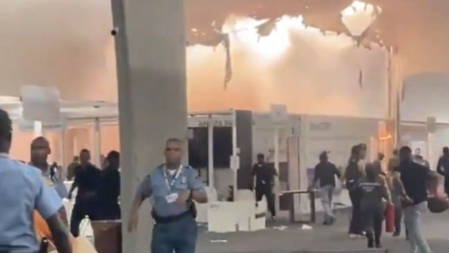 Interior do Pavilhão dos Países da COP30 tomado por fumaça, com teto danificado e pessoas sendo evacuadas por agentes de segurança após incêndio.