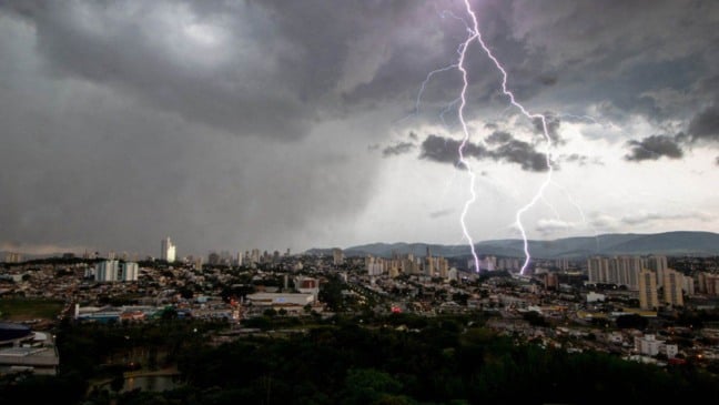 Raio atinge a cidade de Jundiaí durante uma forte tempestade, com nuvens escuras e panorama urbano ao fundo.