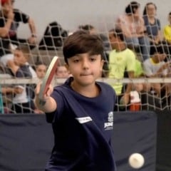 Garoto joga tênis de mesa em quadra coberta durante atividade esportiva em Jundiaí, com público assistindo ao fundo.