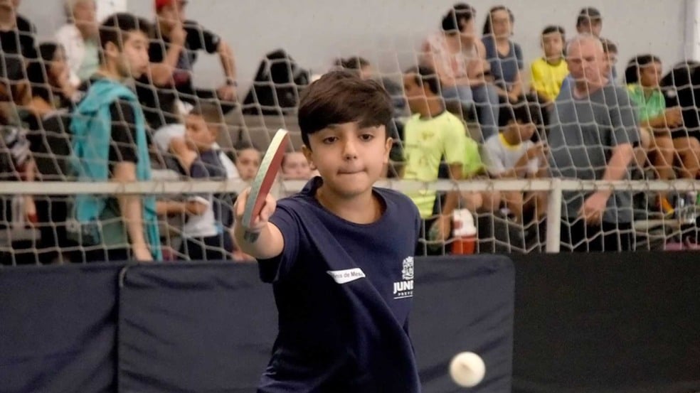 Garoto joga tênis de mesa em quadra coberta durante atividade esportiva em Jundiaí, com público assistindo ao fundo.