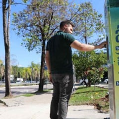 Homem utilizando totem de hidratação da DAE na Avenida Nove de Julho, com árvores, via urbana e céu azul ao fundo.
