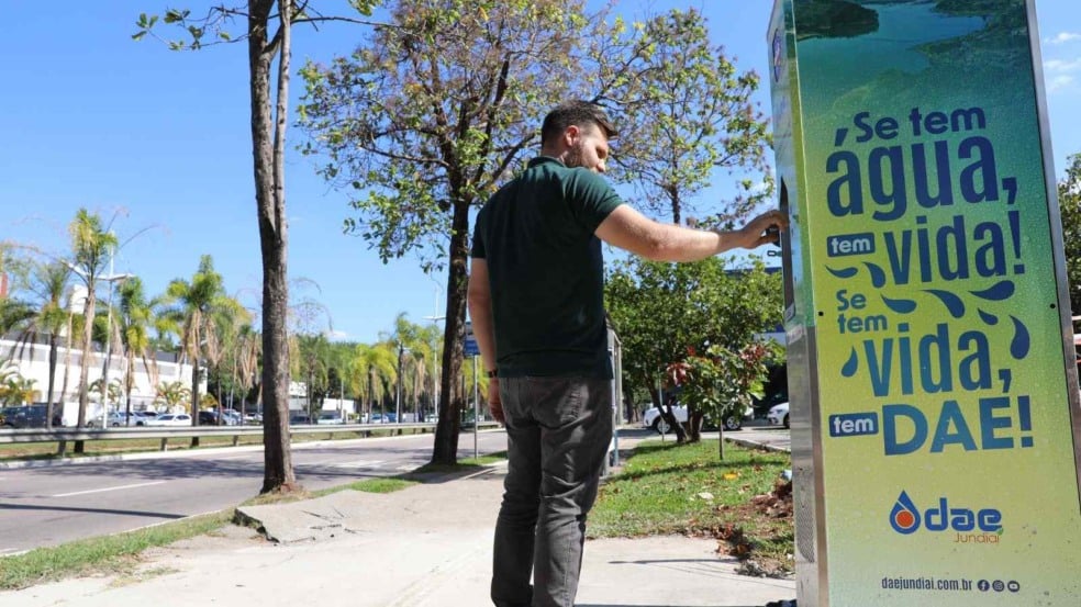 Homem utilizando totem de hidratação da DAE na Avenida Nove de Julho, com árvores, via urbana e céu azul ao fundo. Homem utilizando totem de hidratação da DAE na Avenida Nove de Julho, com árvores, via urbana e céu azul ao fundo.