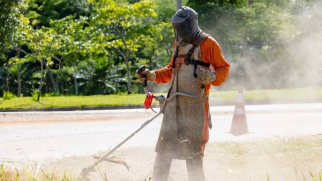 Trabalhador faz roçagem de mato em área pública de Jundiaí, usando equipamento de proteção e máquina motorizada, durante ação de zeladoria urbana.