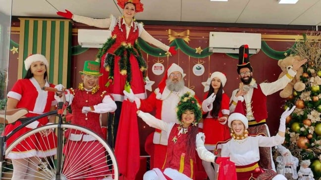 Artistas com figurinos natalinos, incluindo Papai Noel, elfos e personagens circenses, posam em espaço decorado do Natal Luz em Jundiaí.